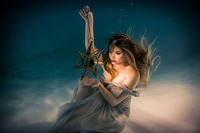 Girl underwater holding a gold and glass star from a metal string.  She is wearing a thin sheet and her hair is flowing in the water.